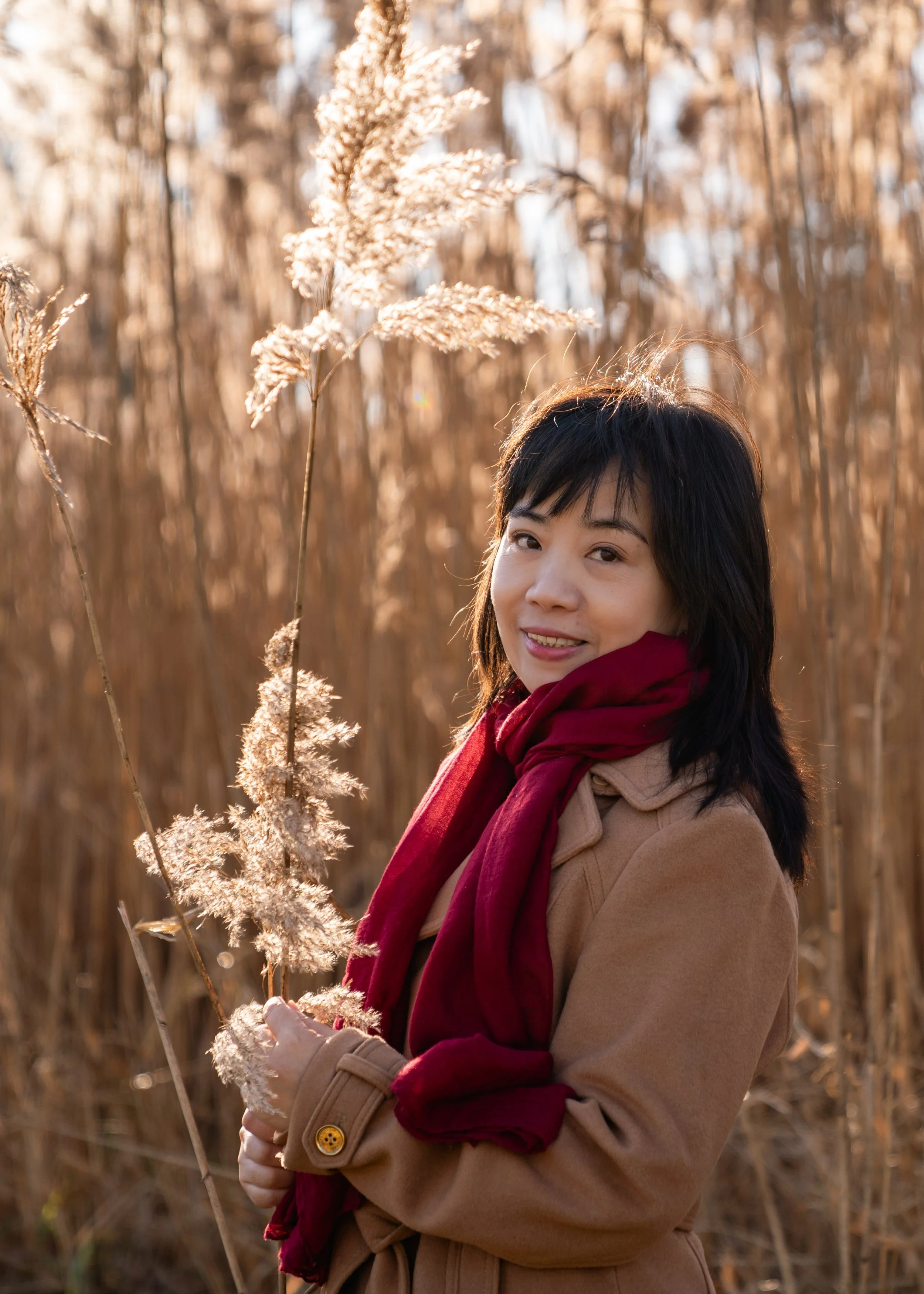 A woman standing among tall dried grasses, wearing a red scarf and smiling gently