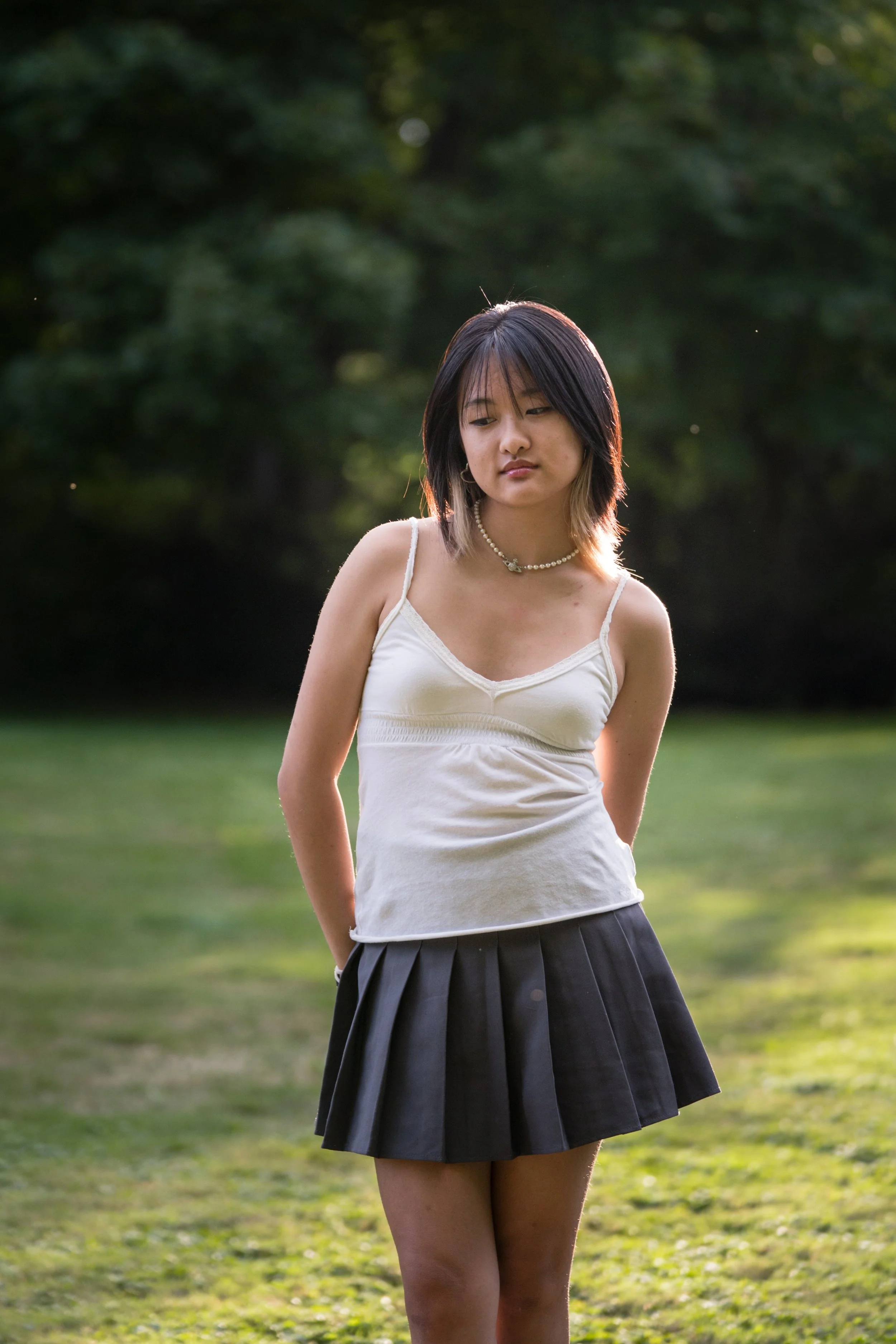 A young woman standing outdoors on a grassy field with trees in the background, wearing a white spaghetti strap top, a black pleated skirt, and a pearl necklace.