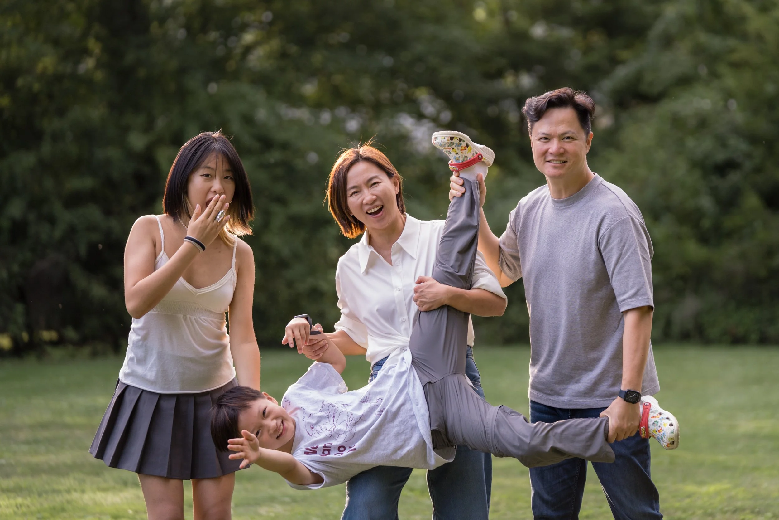 Group of five people, four adults and one child, outdoors in a park. They are smiling and holding the child horizontally by his ankles, lifting him in the air.
