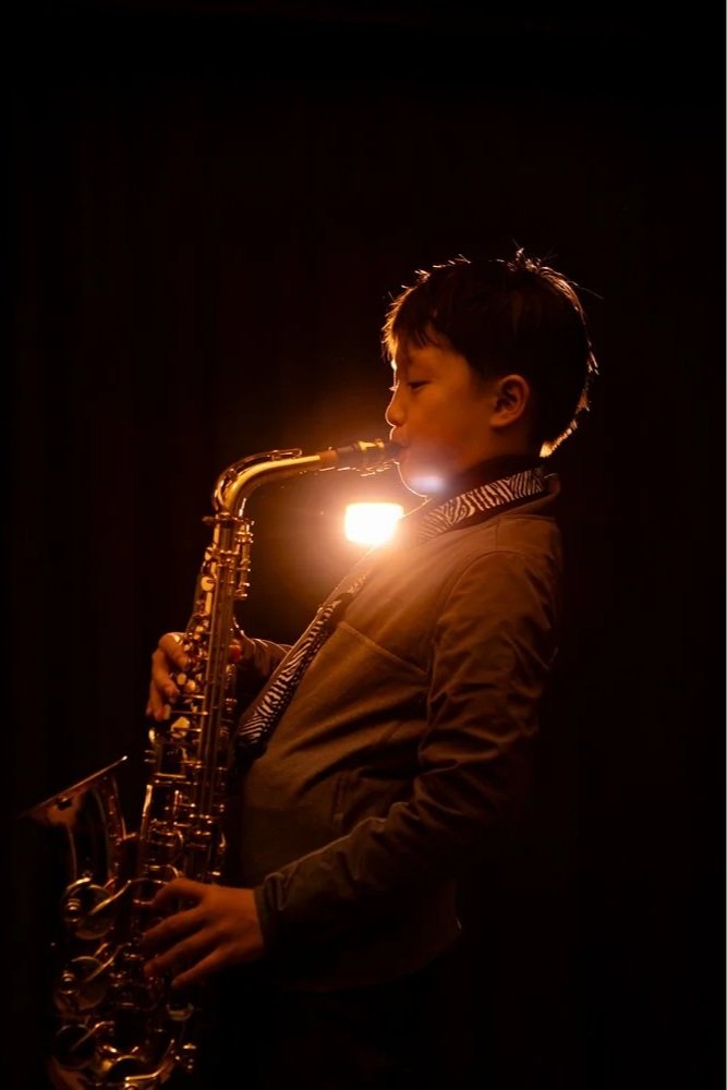 A young boy playing a saxophone in a dark setting with a bright light behind him, creating a silhouette effect.