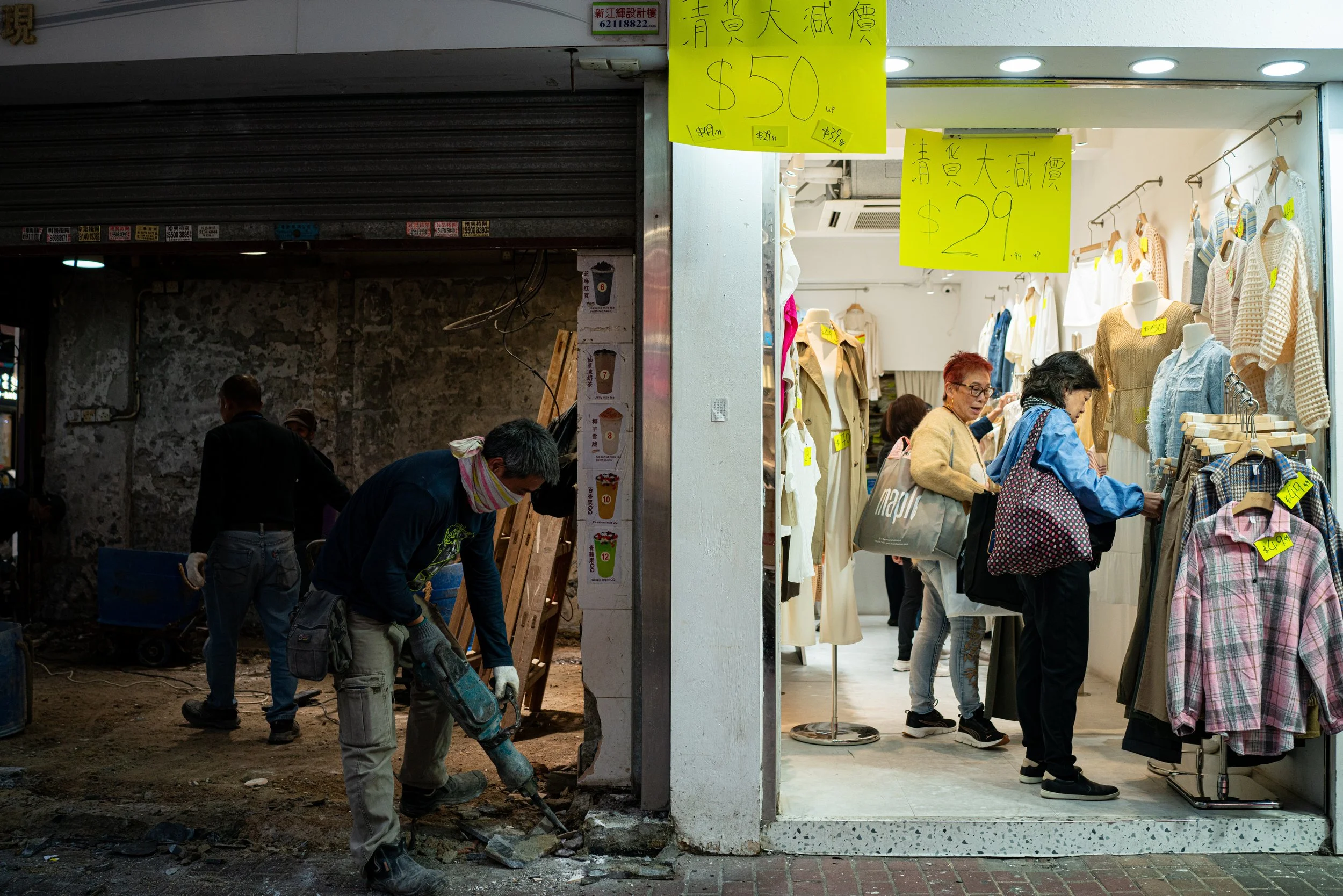 Workers breaking concrete beside an open clothing shop on a city street