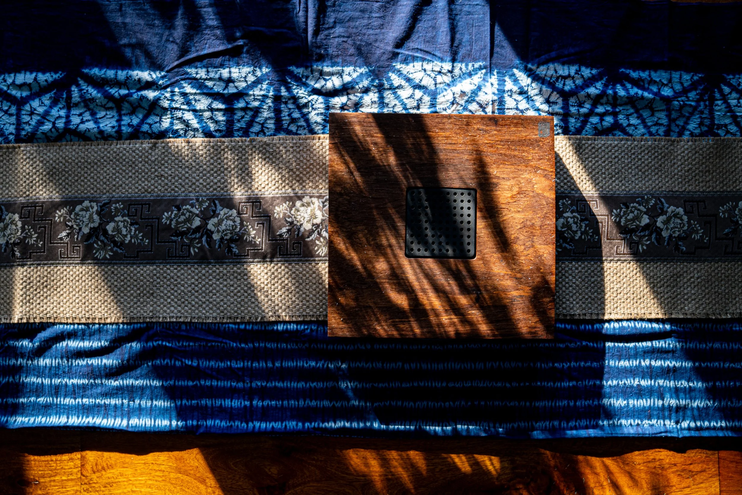 A wooden table with a black square object, possibly a electronic device, placed on top of it. The table is set on decorative fabrics with floral and geometric patterns, illuminated by natural light creating shadows.