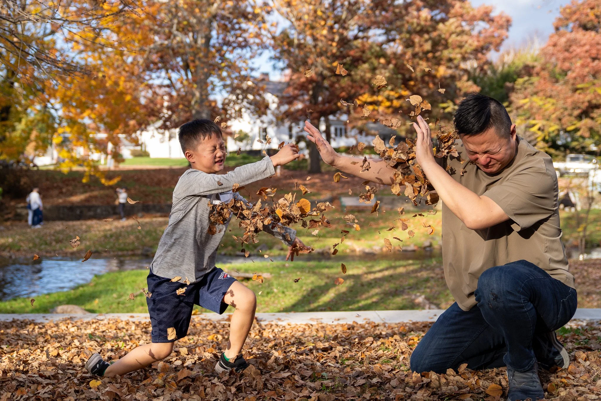 A boy and an adult man playing in a park during fall, throwing and covering each other with dry leaves. The boy is running while the man kneels with his hands up, trying to block the leaves. There are autumn trees and a river in the background.