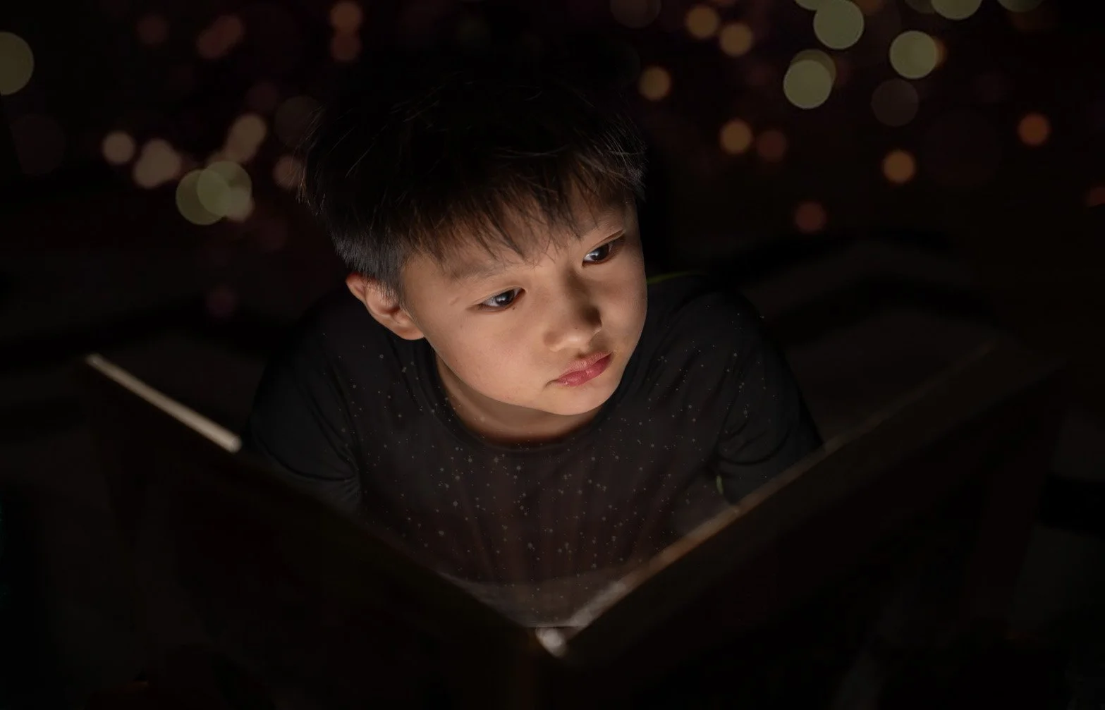 A young boy with dark hair looking at a glowing device, with a dark background featuring blurred lights.