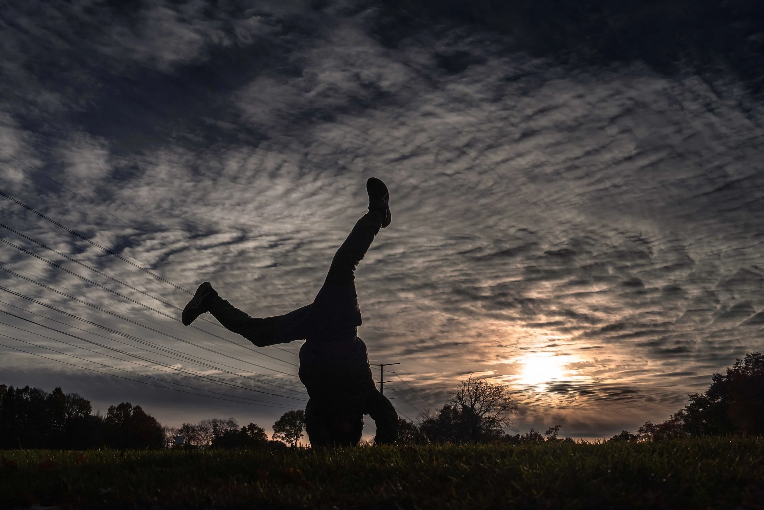 A silhouetted figure balancing upside down on grass at sunset beneath textured clouds