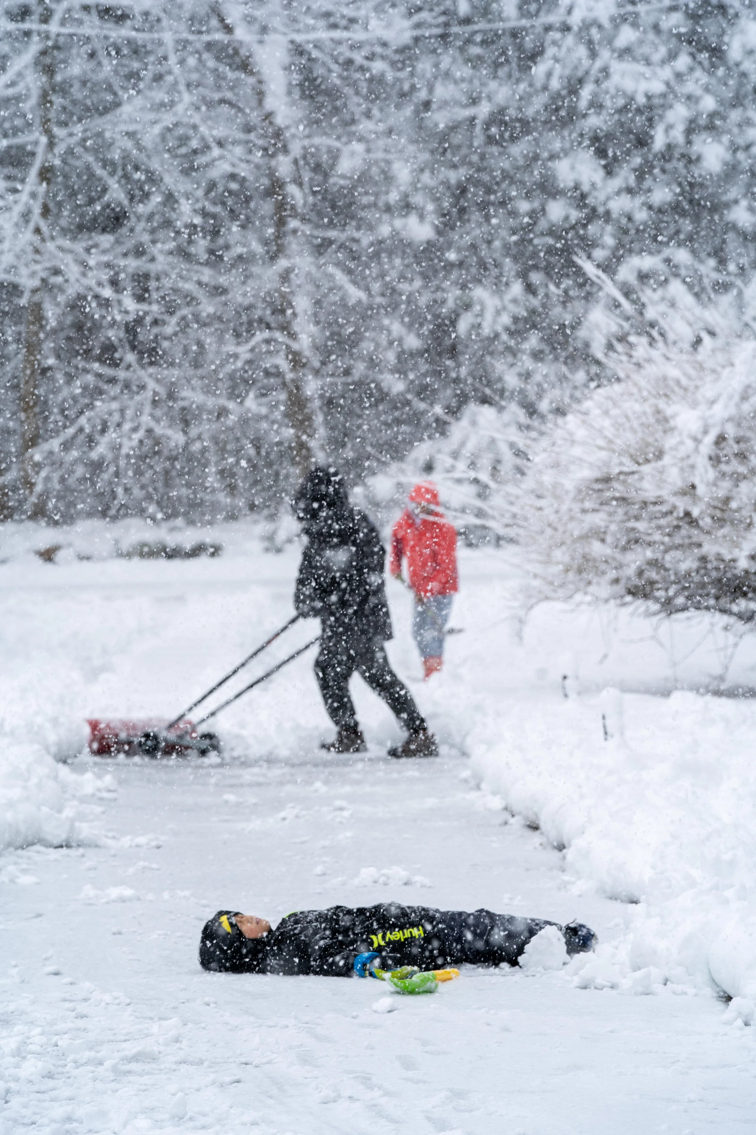 Person lying on snow-covered ground in winter, two people shoveling snow nearby in a snowy outdoor setting.