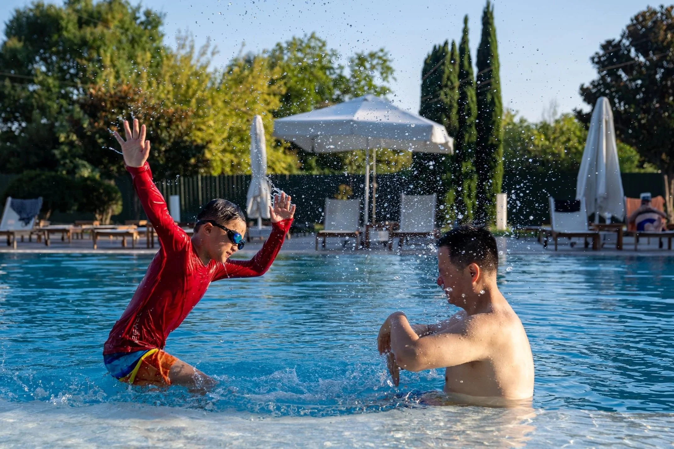 Two boys playing and splashing water in a pool on a sunny day, with pool chairs and umbrellas in the background.