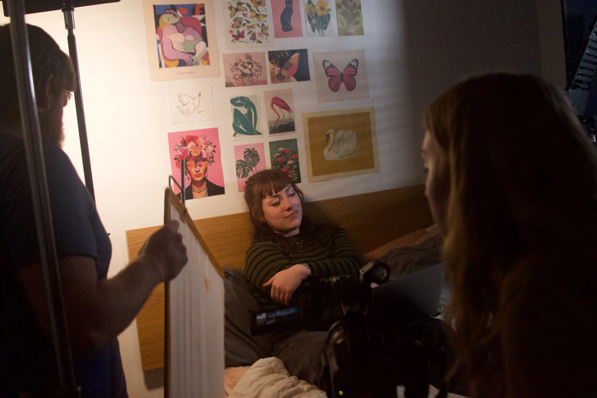 A woman lying in bed with her eyes closed and arms crossed, with two women filming her in a dimly lit room. The wall behind her is decorated with colorful artwork, including floral patterns, a butterfly, and a woman with a floral headdress.