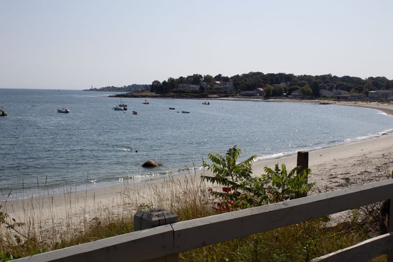 A peaceful beach scene with calm water, small boats floating, sandy shoreline, and houses in the background, viewed from a deck with railing and some greenery in the foreground.