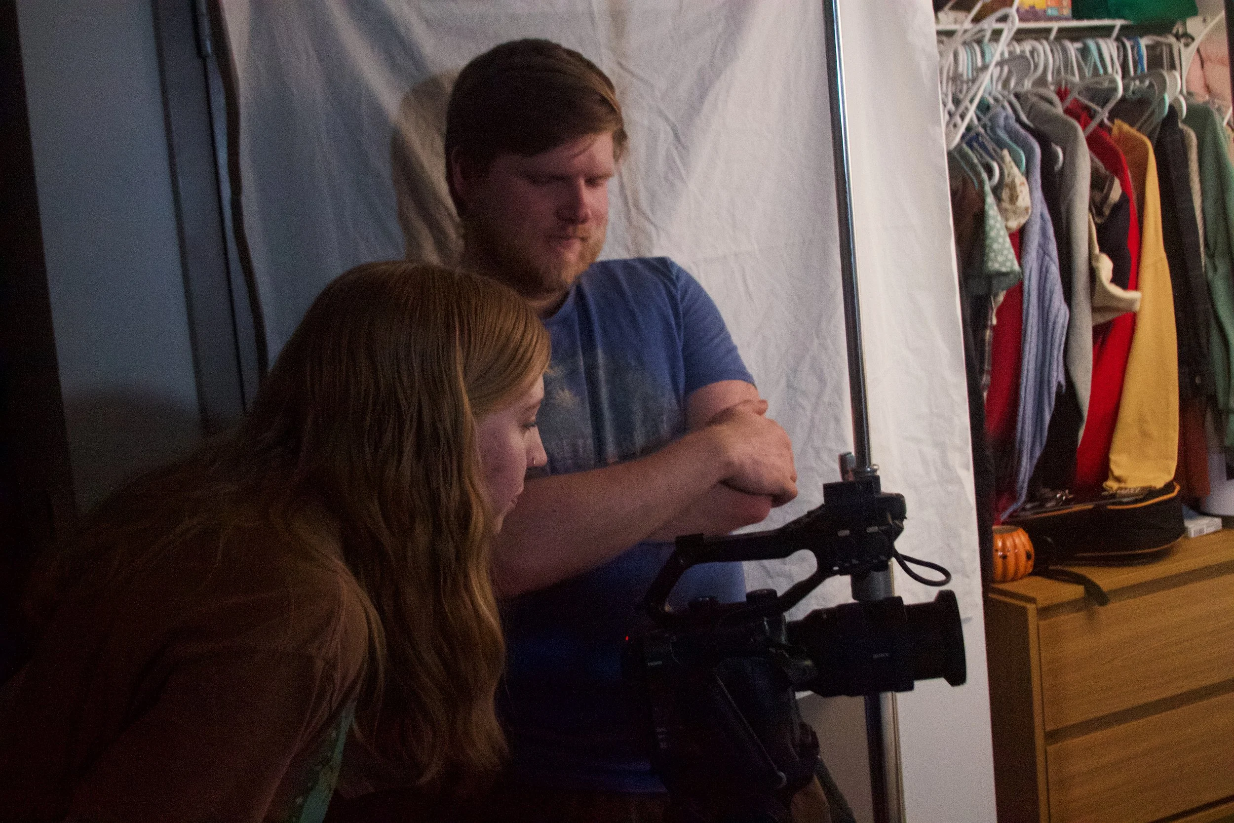 Two people setting up a camera in front of a white backdrop, with a clothing rack filled with colorful clothes in the background.