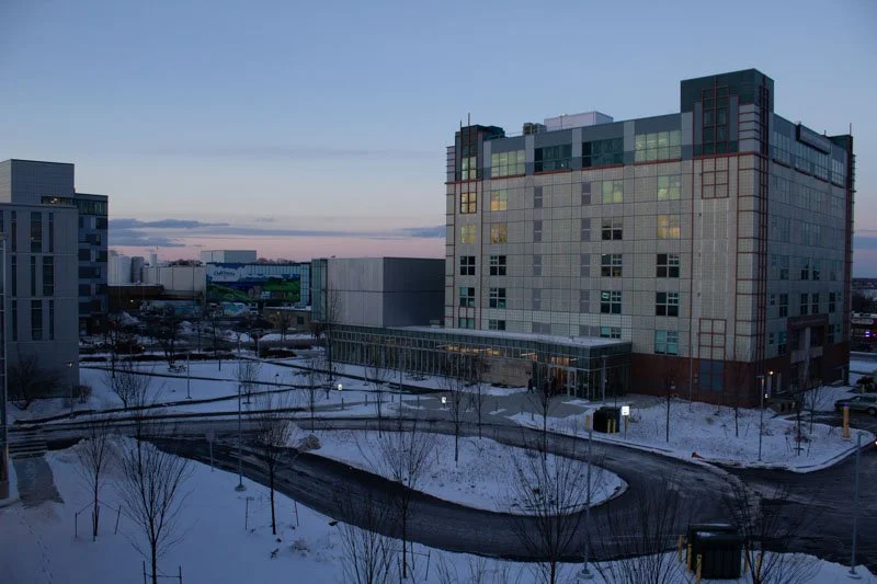 A large modern building in a snowy cityscape during dusk, with other buildings and leafless trees visible, and a winding parking lot.