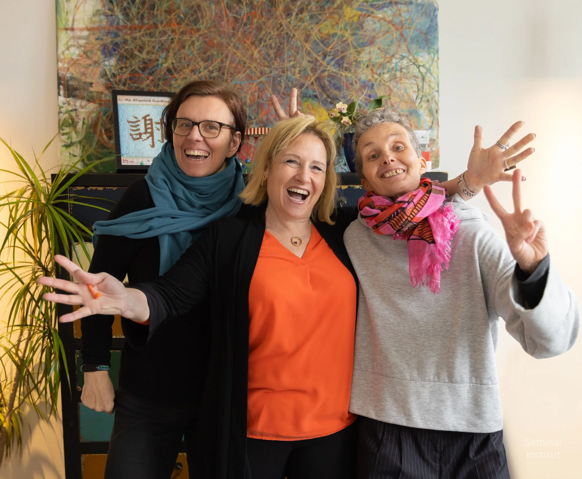 Drei fröhliche Menschen lachen und zeigen Peace-Zeichen im Büro, mit bunter Wandkunst im Hintergrund und einer Pflanze auf der linken Seite.