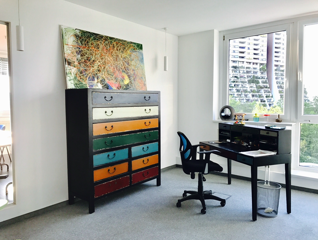 Ein modernes Arbeitszimmer mit einem schwarzen Schreibtisch, einem schwarzen Bürostuhl, einer Kommode mit bunten Schubladen und einem großen Fenster mit Blick auf Hochhäuser.