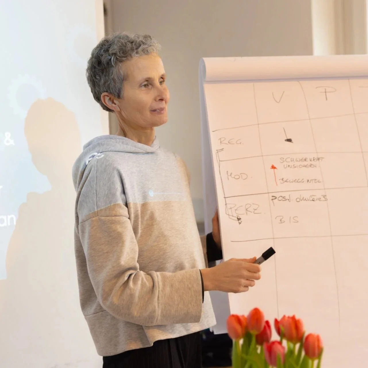 Eine Frau mit kurz grau Locken in einem grauen Hoodie präsentiert eine Flipchart während eines Unterrichts oder einer Präsentation, mit rotem Tulpenstrauß im Vordergrund