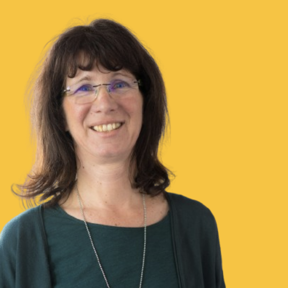 Porträt einer lachenden Frau mit braunen Haaren, Brille und dunkler Kleidung vor einem gelben Hintergrund.