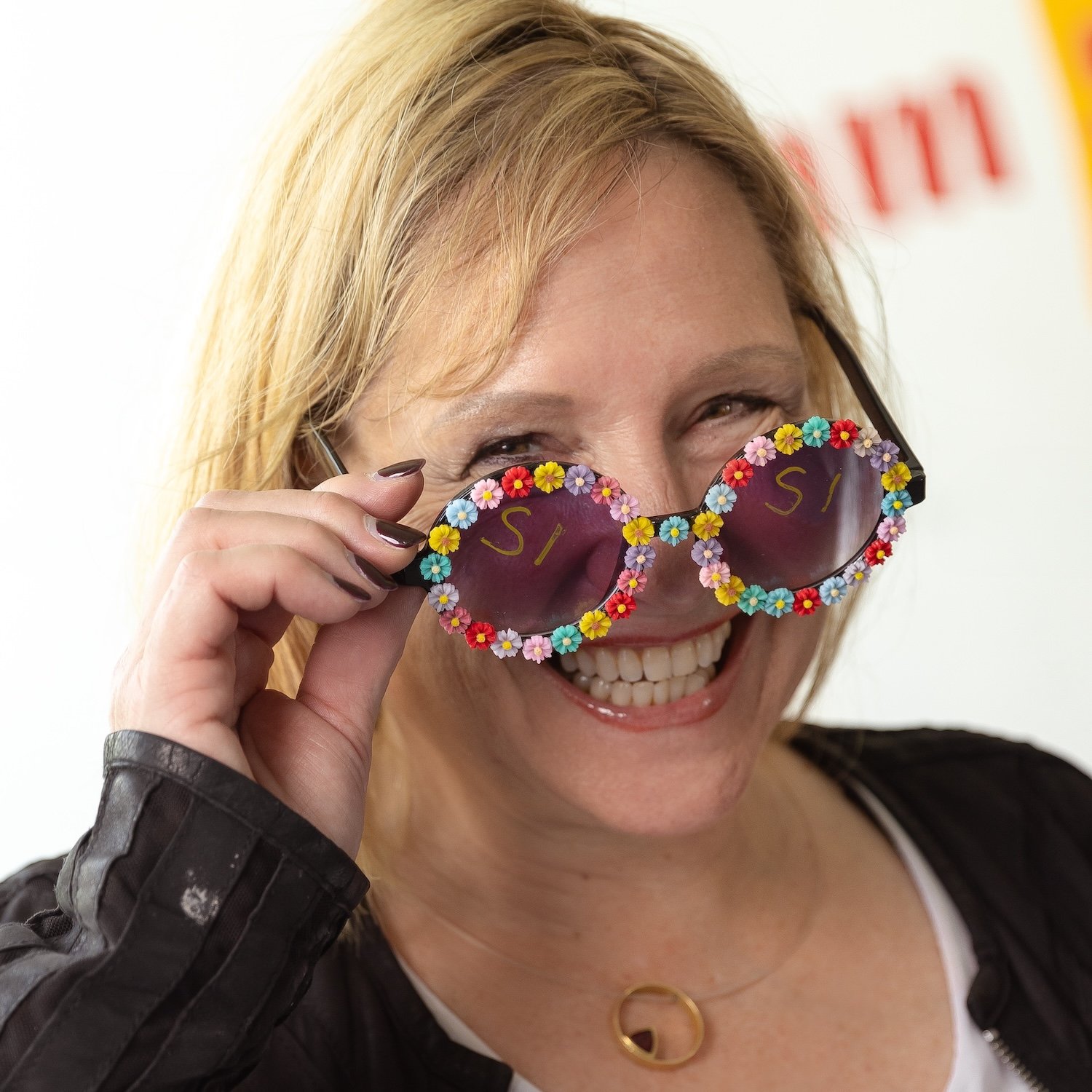 Eine lachende Frau mit blonden Haaren, die bunte, blumenumrahmte Sonnenbrille trägt.