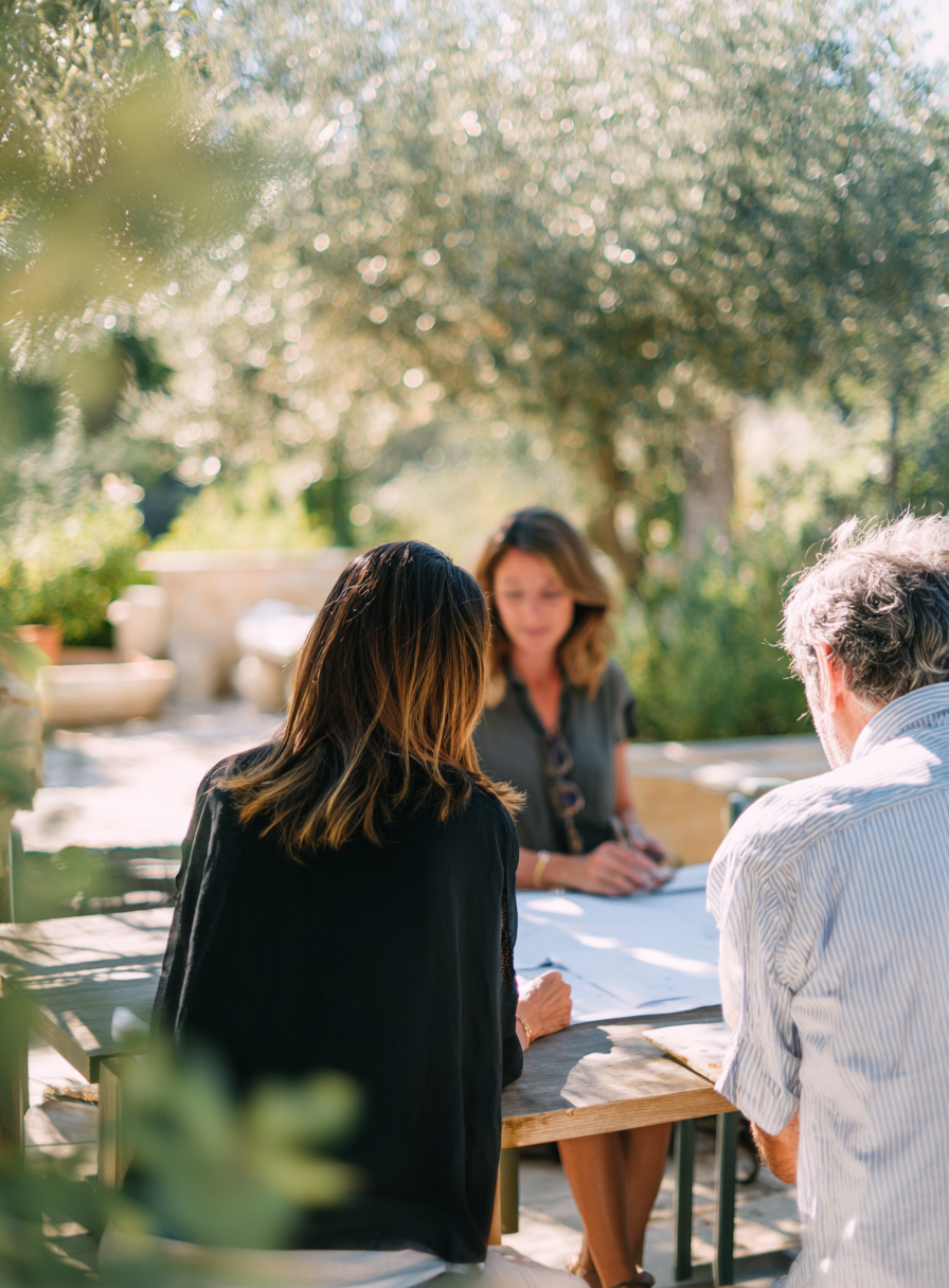 Trois personnes assises autour d'une table en extérieur, en train de discuter ou de signer des documents, sous un arbre avec beaucoup de lumière naturelle.