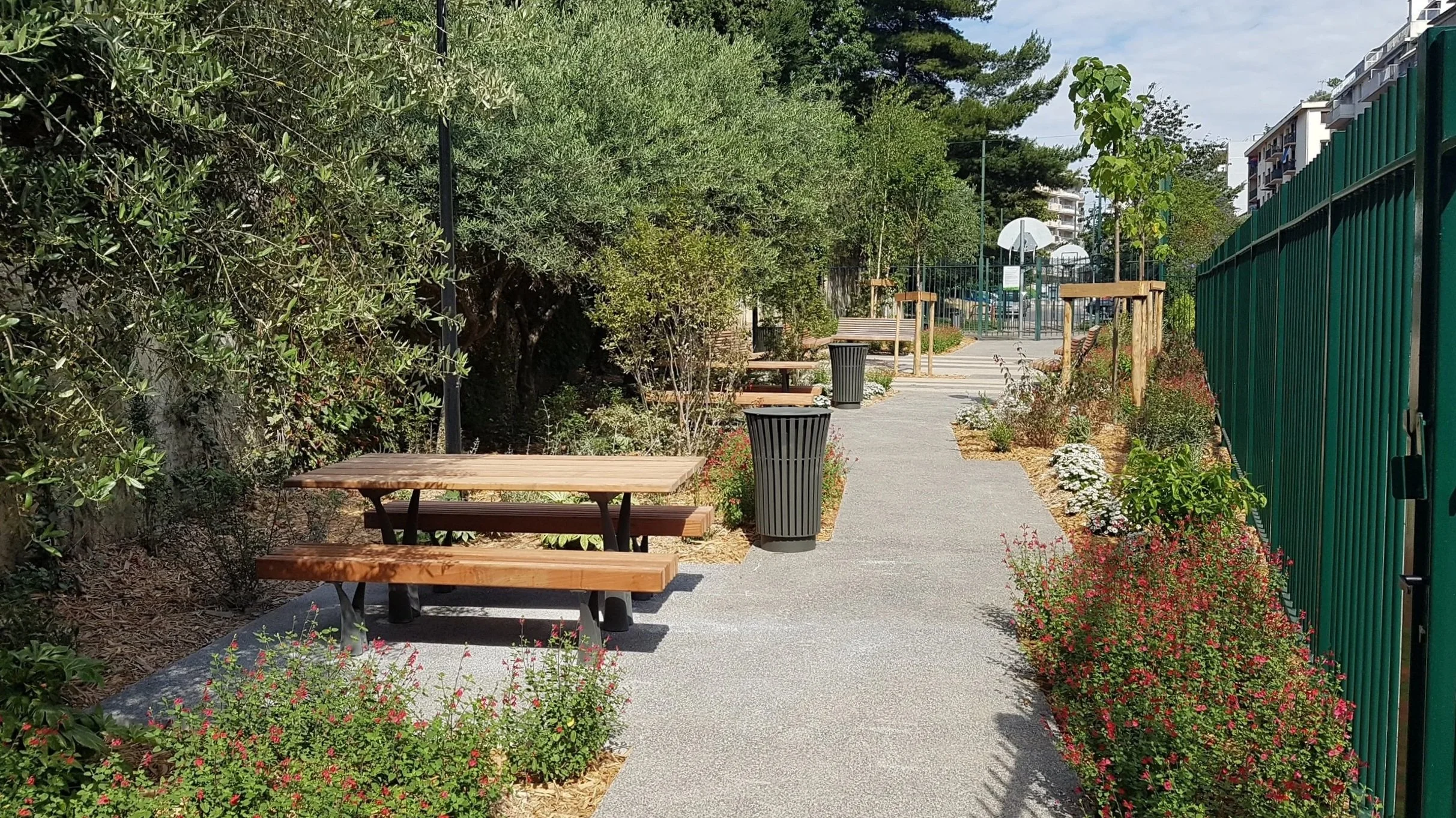 Sentier de parc avec bancs en bois, poubelles, plants et arbustes, clôture verte et terrains de basketball au fond.