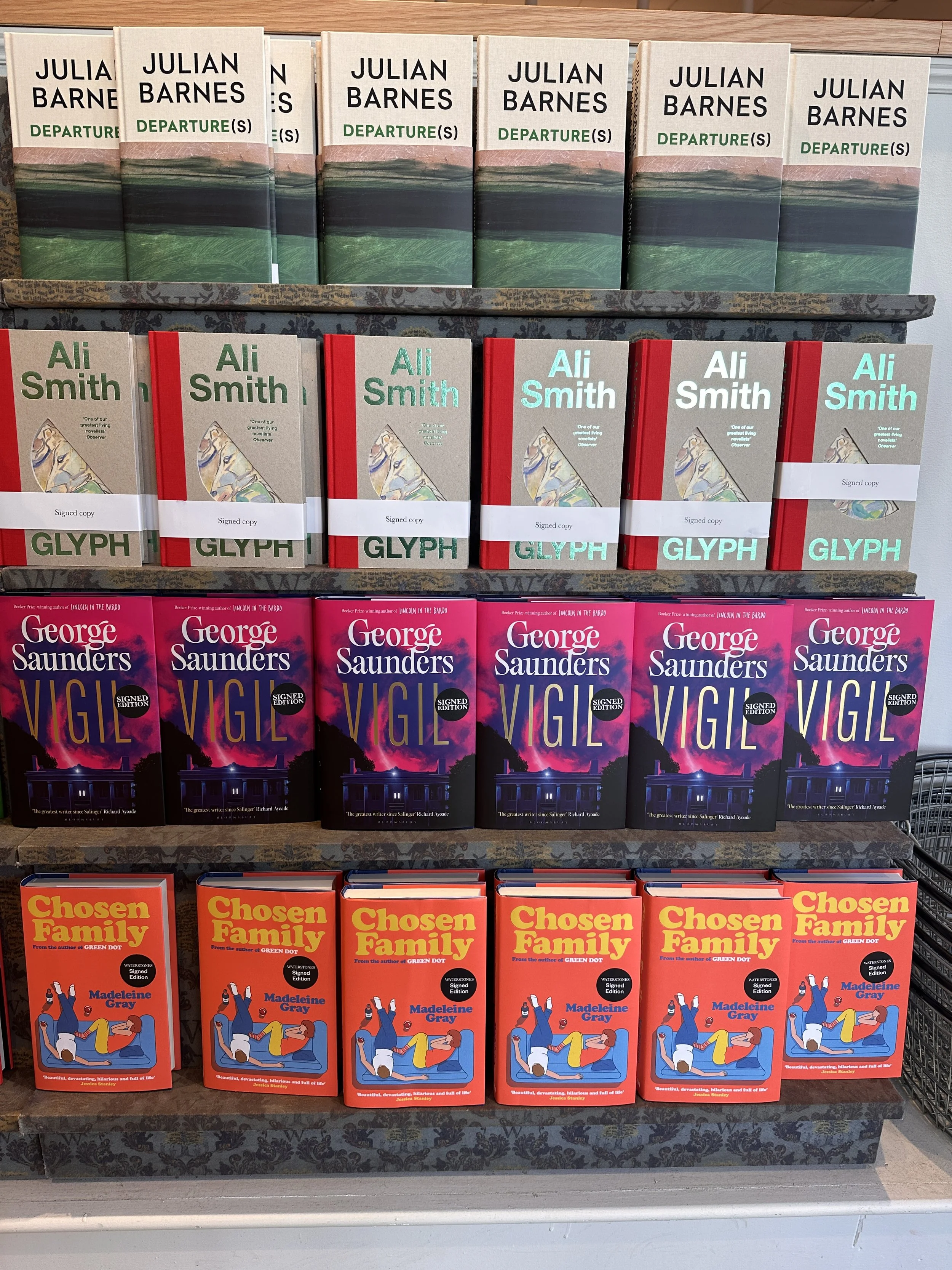 Books arranged on shelves with four different titles. The top shelf has multiple copies of 'Departure' by Julian Barnes. The second shelf has copies of 'Glyph' by Ali Smith, with some marked as signed copies. The third shelf has several copies of 'Vi
