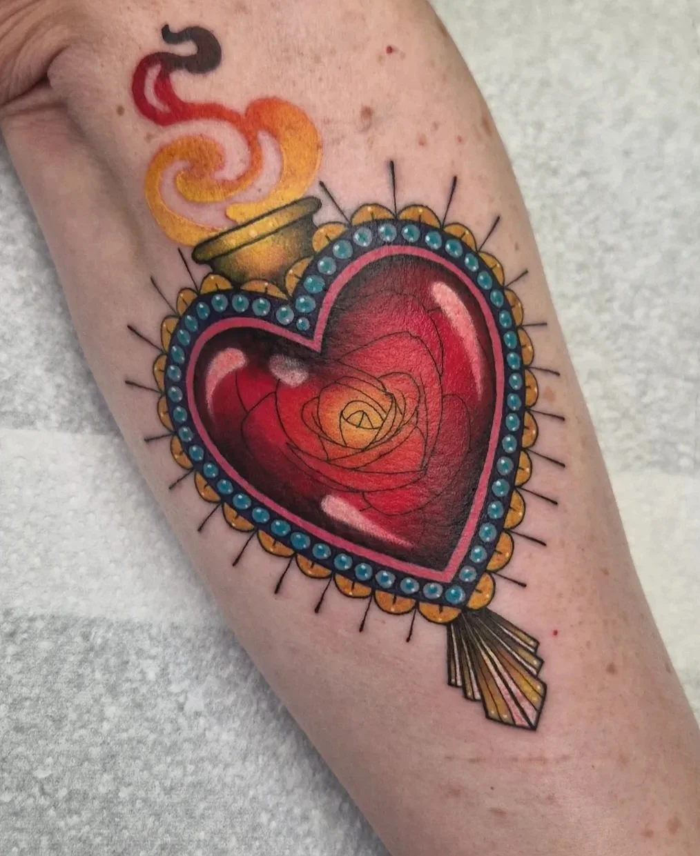 Full colour tattoo of a shiny, red sacred heart with a rose inside, surrounded by a beaded border and fine line details. 