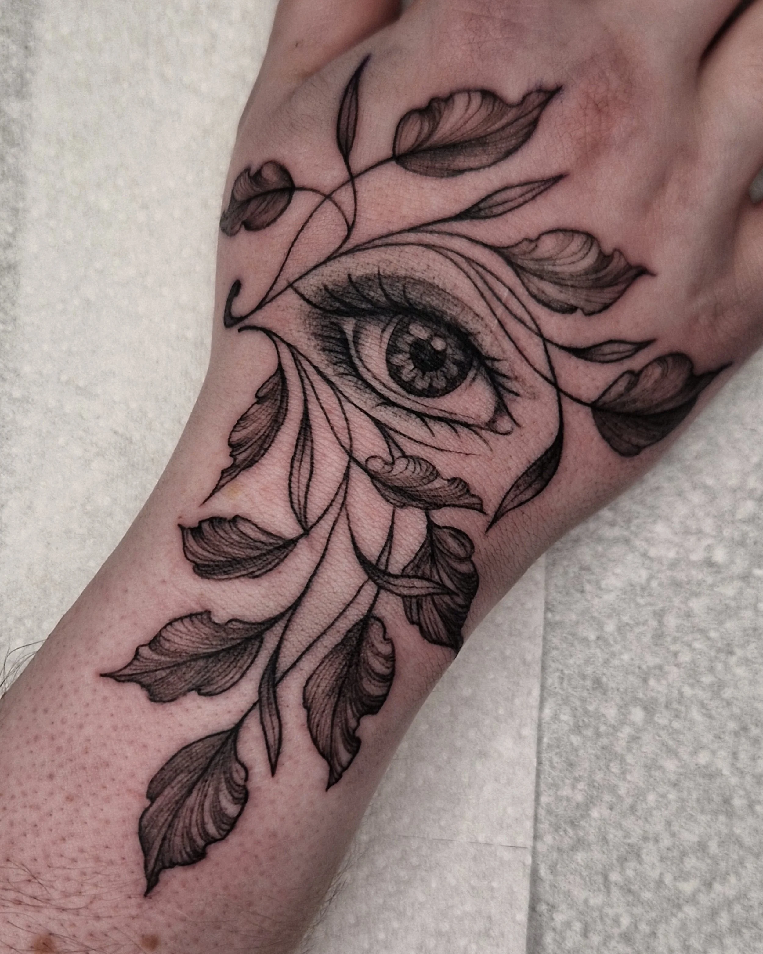 A hand tattoo of a realistic eye with detailed eyelashes and iris, surrounded by leaves.