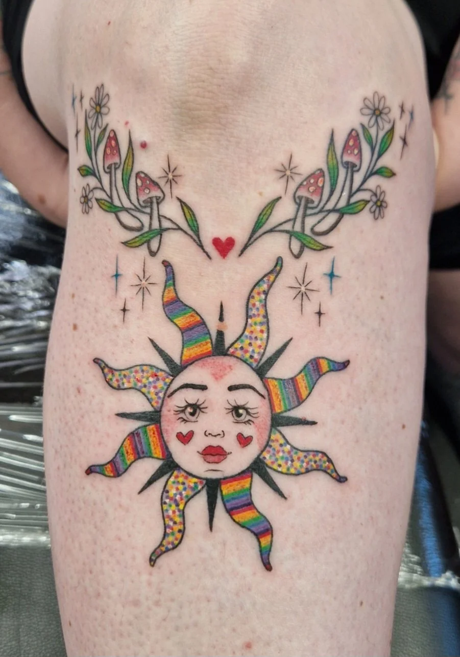 Colorful tattoo of a sun with a face and rainbow-colored rays, surrounded by stars, a  hearts, and vines with pink mushrooms and daisies.