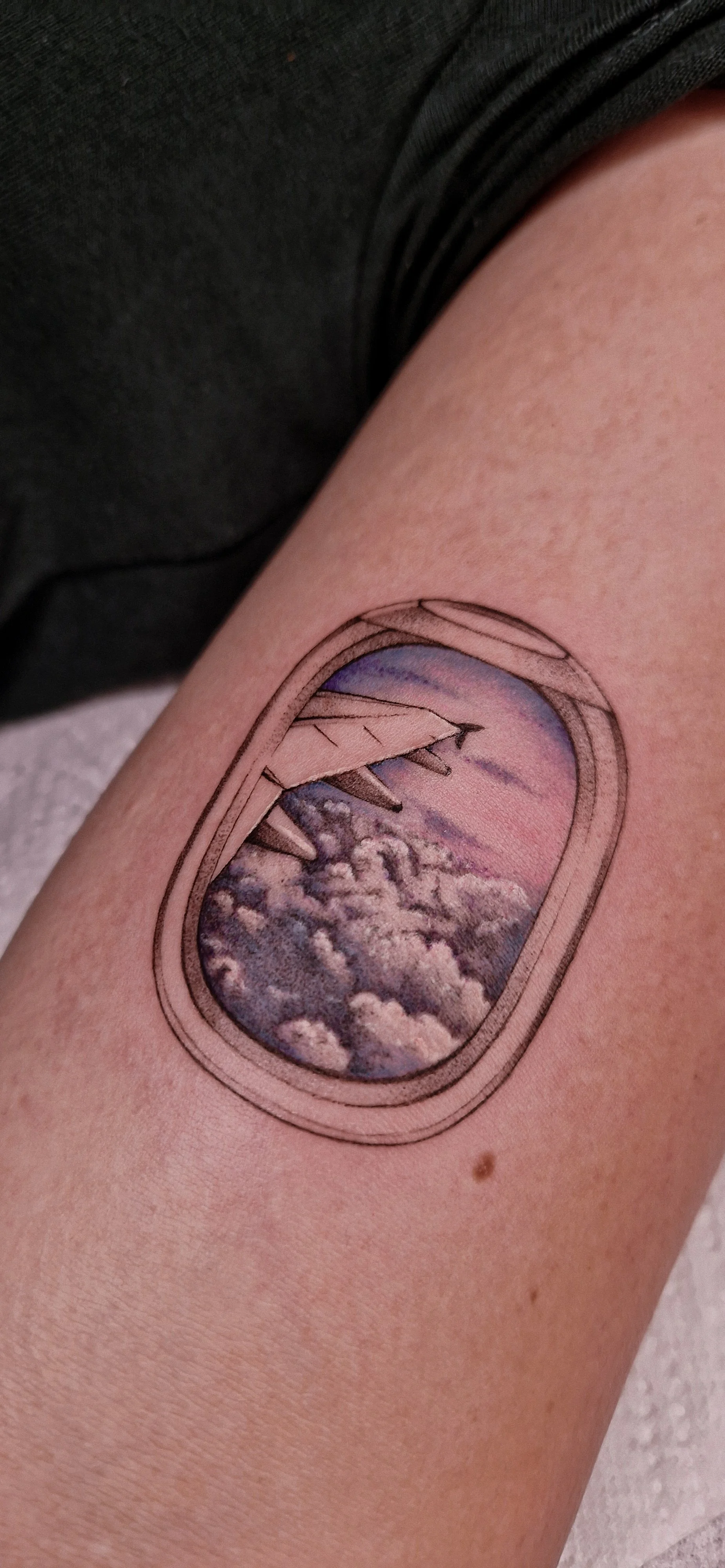Full colour tattoo of an airplane window view showing pink sky and fluffy clouds, with a wing partially visible, on a person's arm.