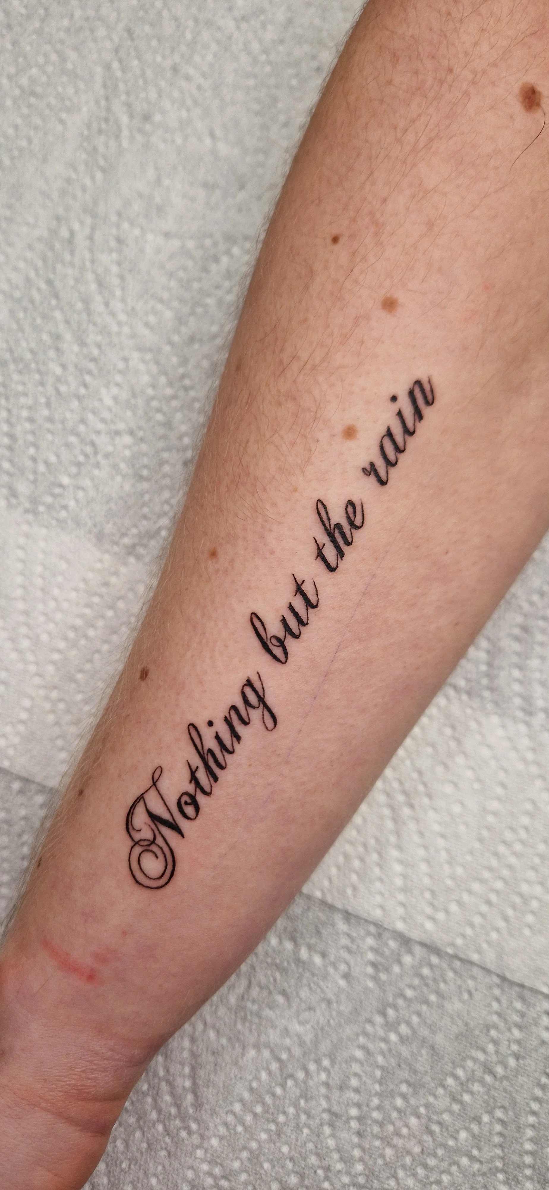 Tattoo on forearm with the phrase 'Nothing but the rain' in black cursive script.