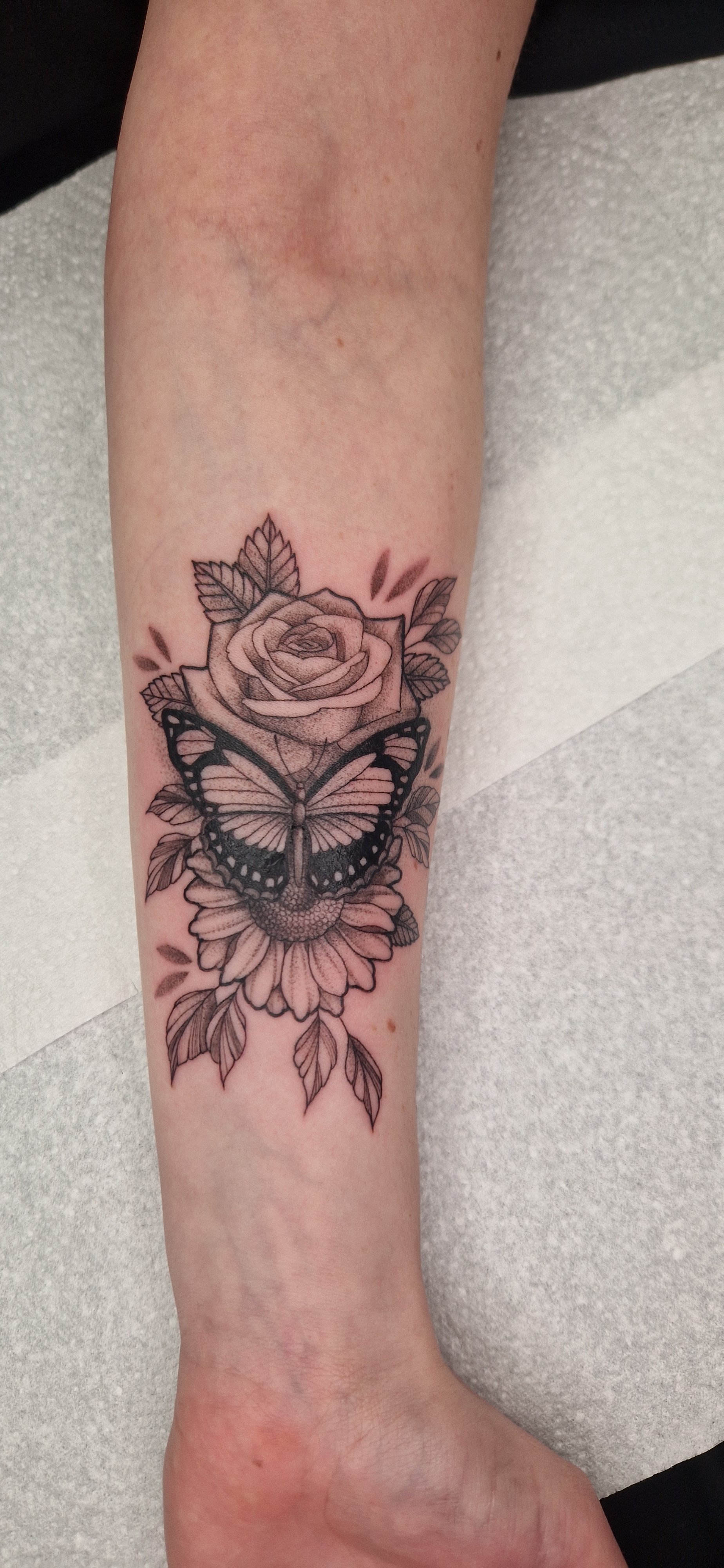 Black and grey tattoo of a butterfly on a rose with leaves and sunflower on a person's arm.