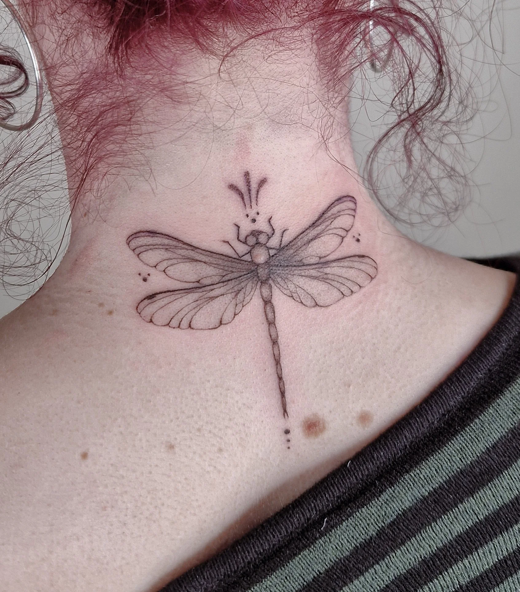 Soft fine line tattoo of a dragonfly in black and grey on the back of the neck.
