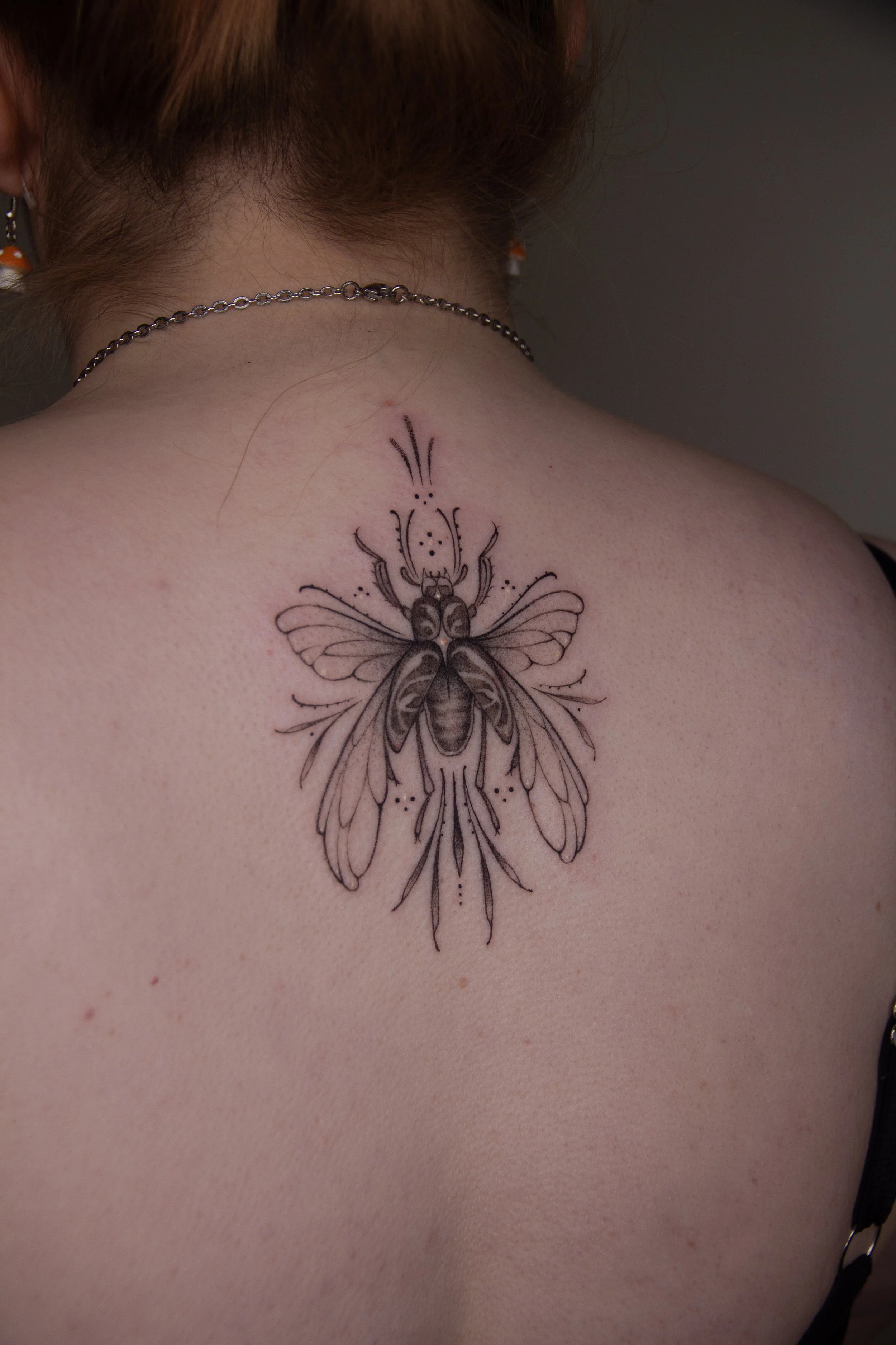 A detailed black and gray tattoo of a beetle with decorative lines and dots on a person's upper back and neck.