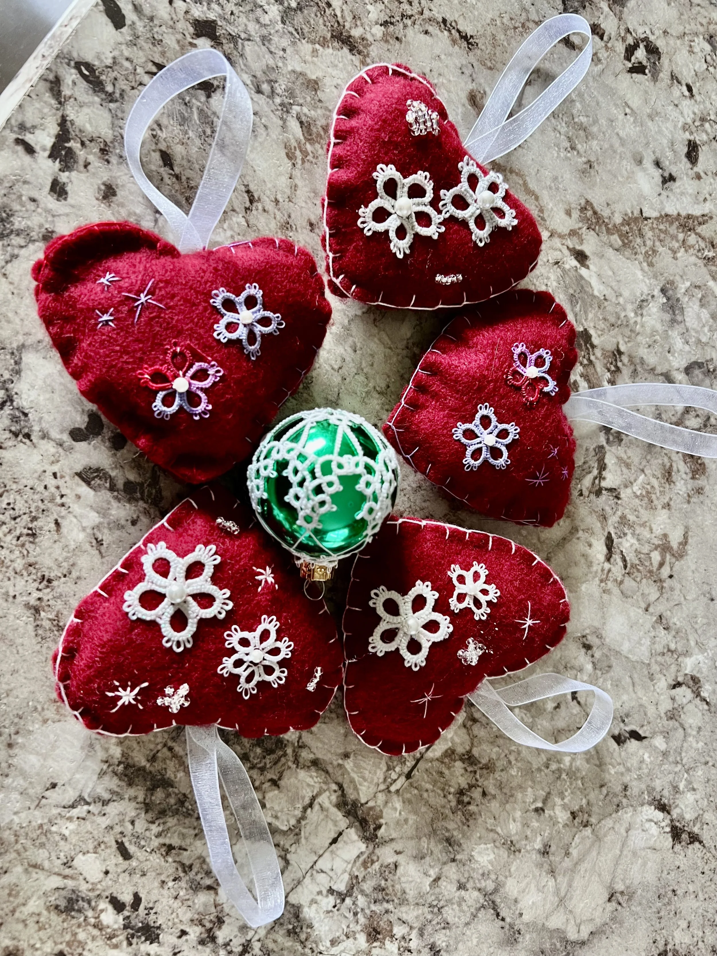 Five red felt heart ornaments with white lace and embroidery, each with a white ribbon for hanging, surrounding a green ornament with a white lace pattern, on a marble surface.