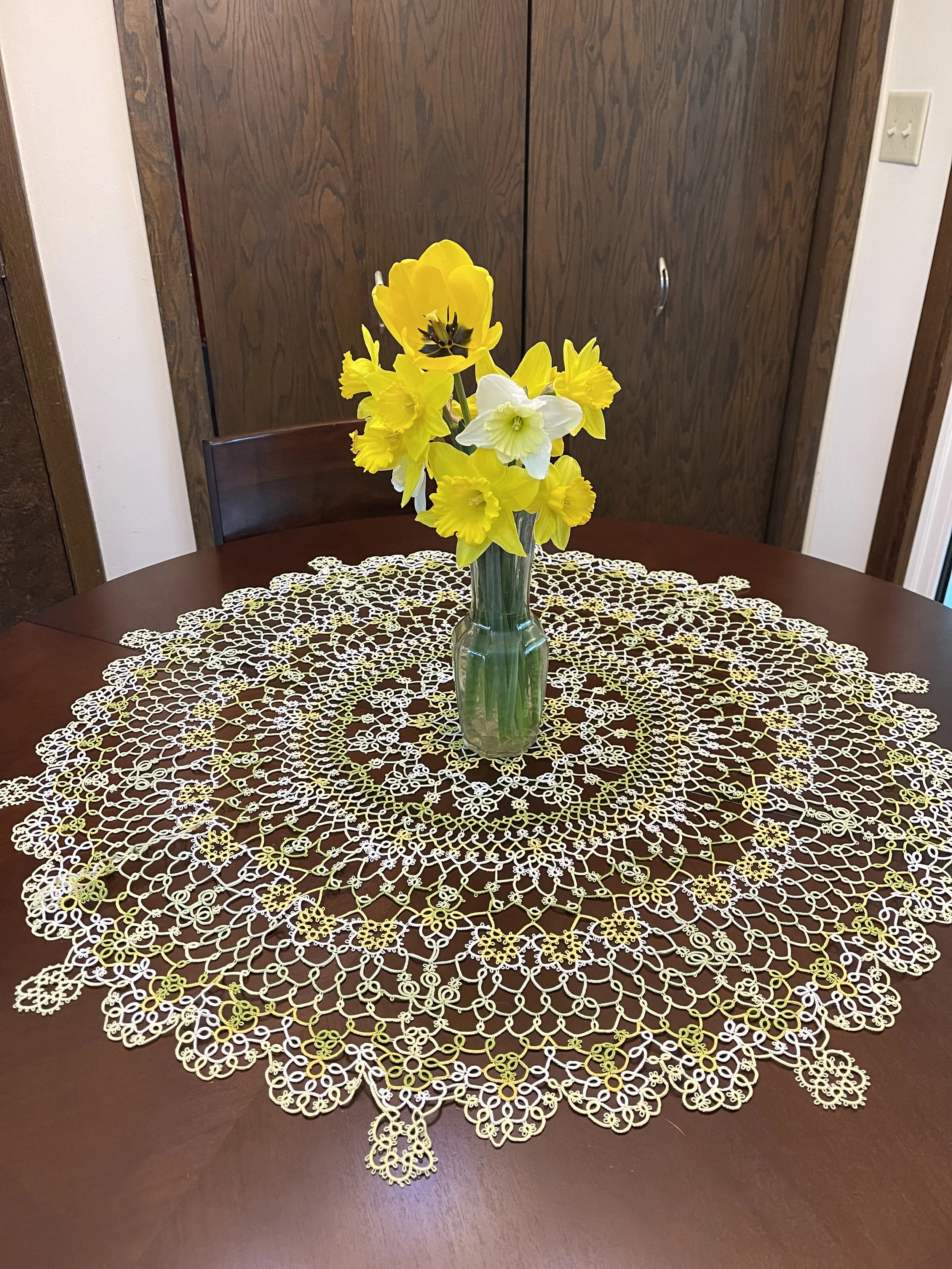A glass vase with yellow and white flowers on a round, lace tablecloth on a wooden table in a dining room.