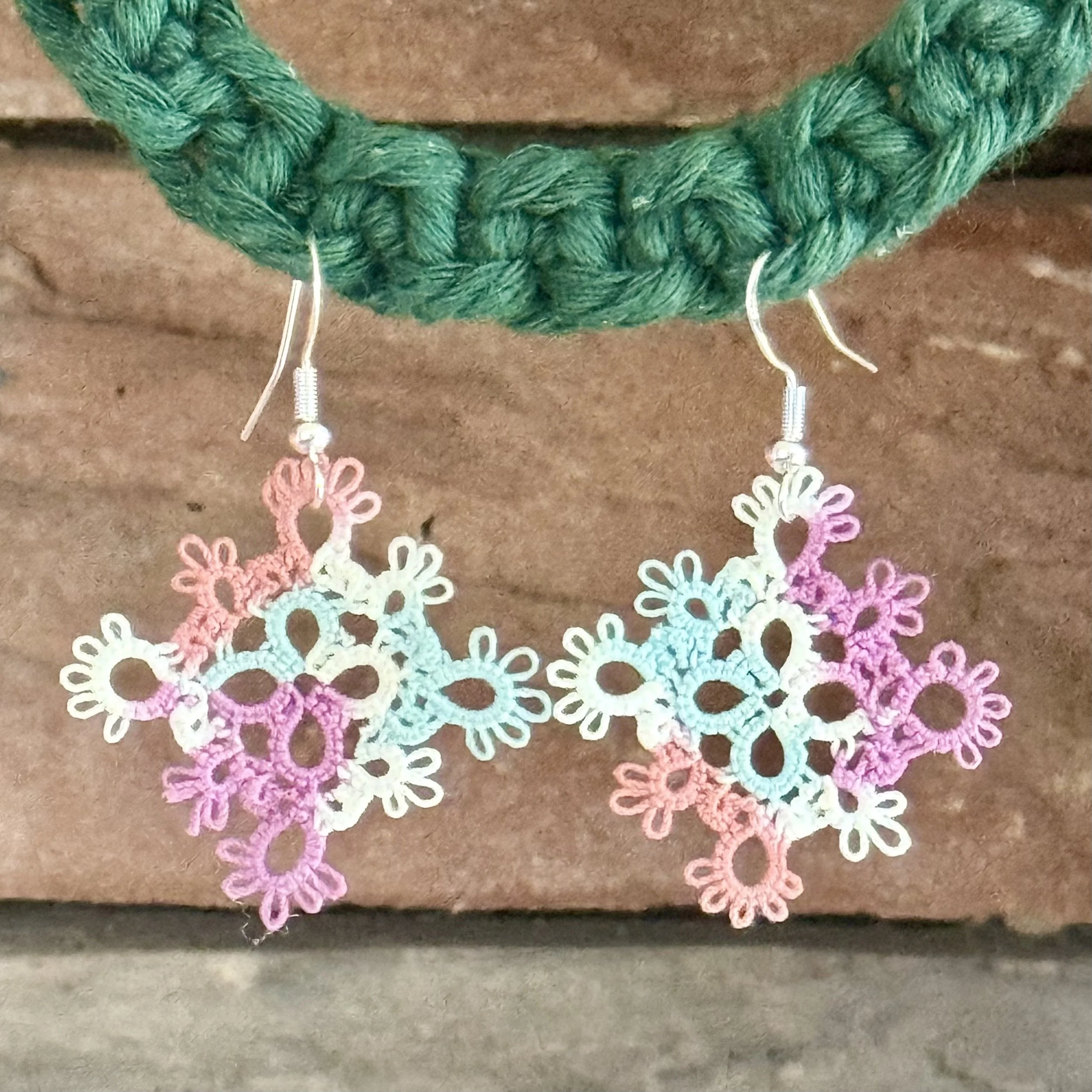 Colorful crochet snowflake earrings hanging from a green woven bracelet against a brown background.