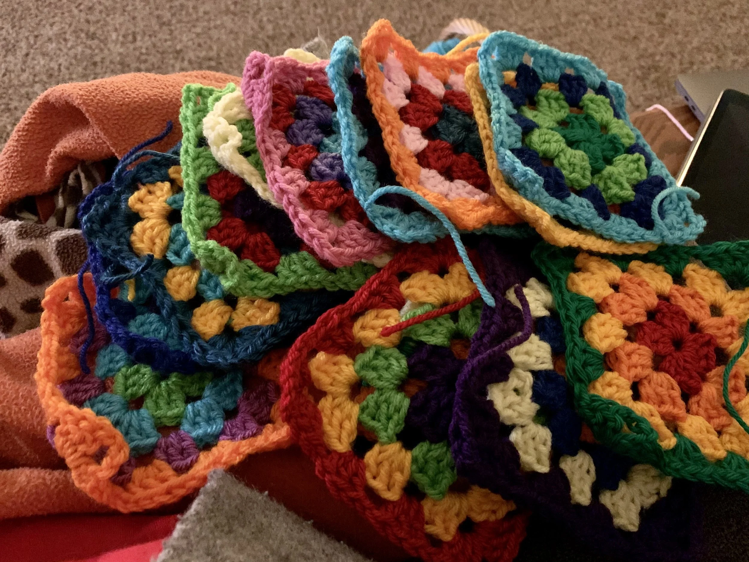 Multiple colorful crocheted granny squares stacked on a surface, with a partly visible phone on the side.