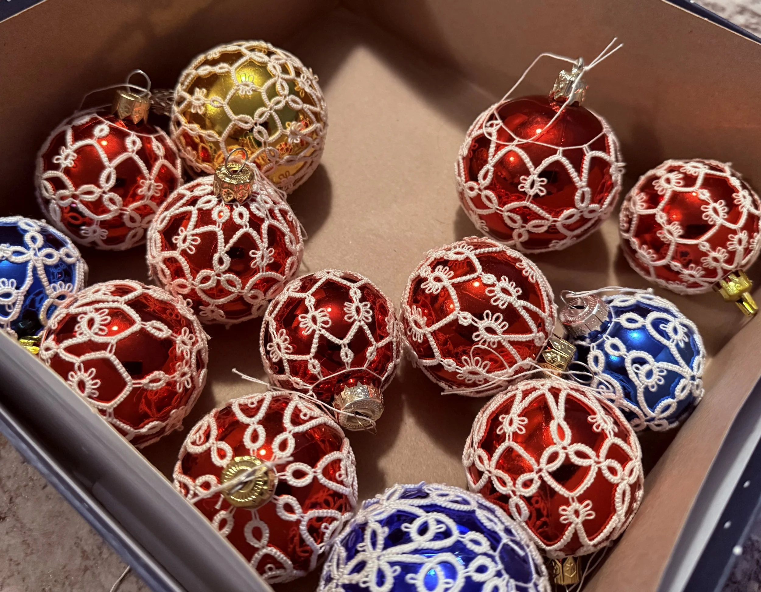 A collection of Christmas ornaments, mostly red with a few blue and gold, decorated with white lace-like string designs, inside a cardboard box.
