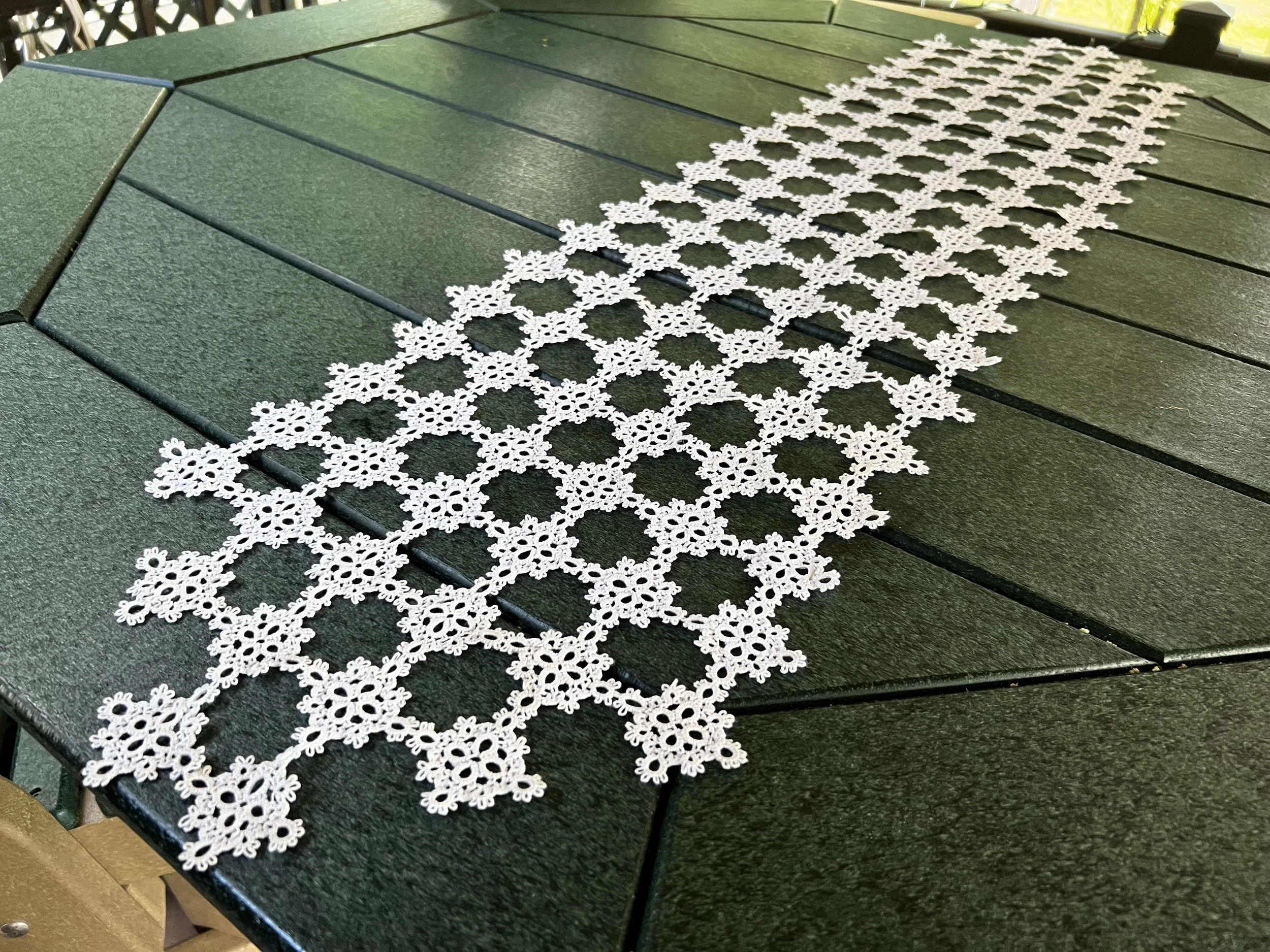 A white crochet table runner with floral patterns laid out on a dark green outdoor table.
