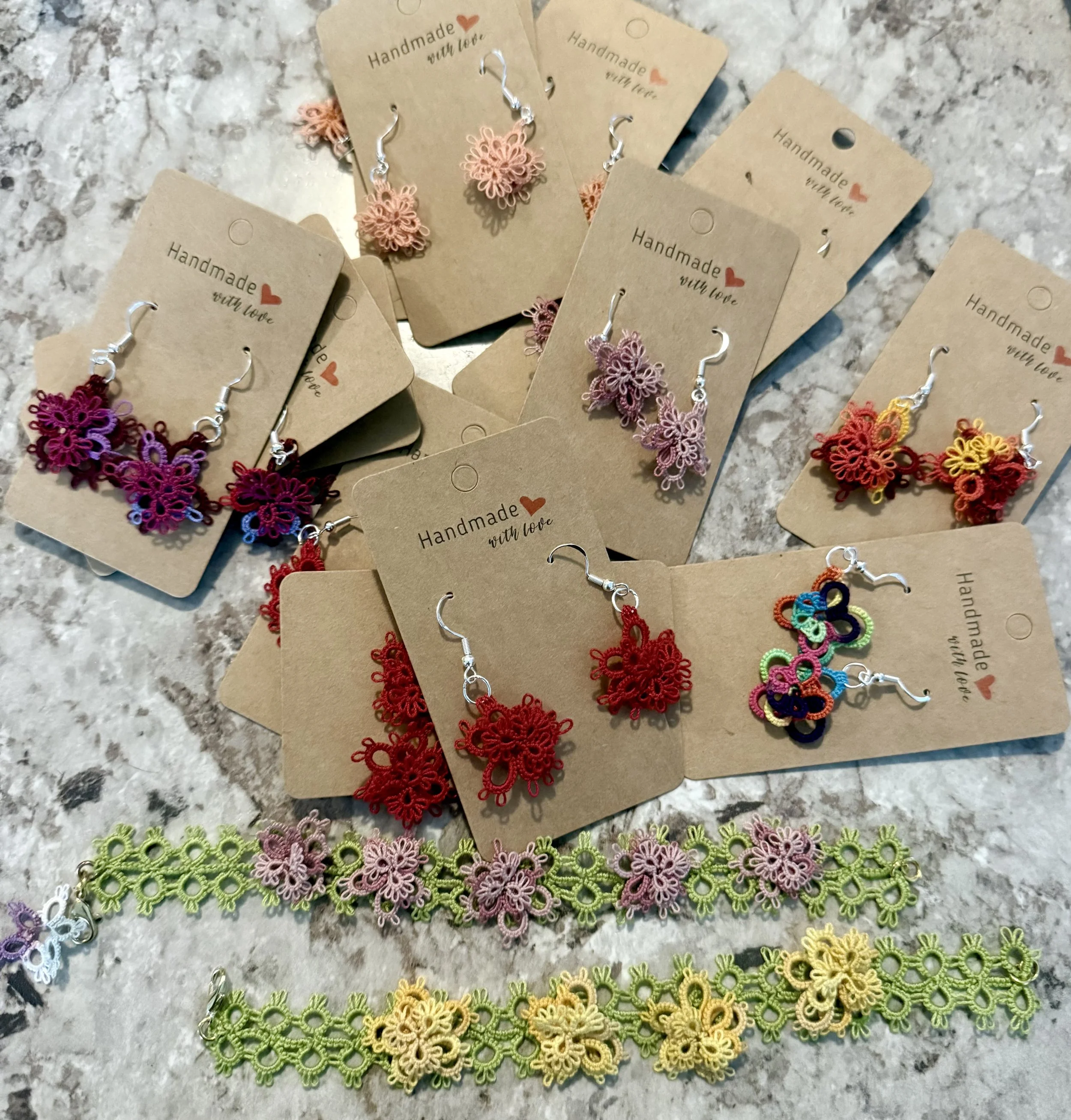 Assorted handmade jewelry including earrings and lace bracelets displayed on brown craft tags labeled 'Handmade with love' on a gray marble surface.