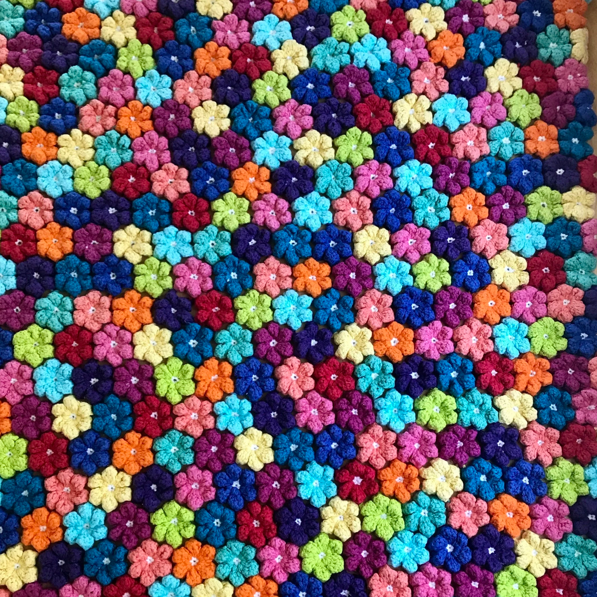 A colorful display of many small, knitted flowers in various shades including pink, blue, purple, green, red, orange, and yellow, arranged in a densely packed pattern.