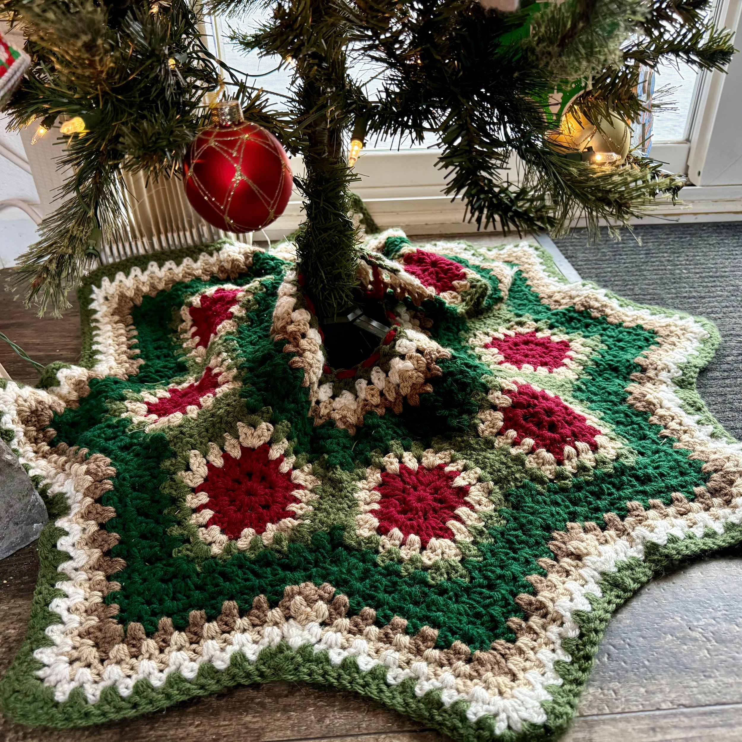 A decorated Christmas tree with red and gold ornaments, standing on a colorful crocheted holiday rug near a window.
