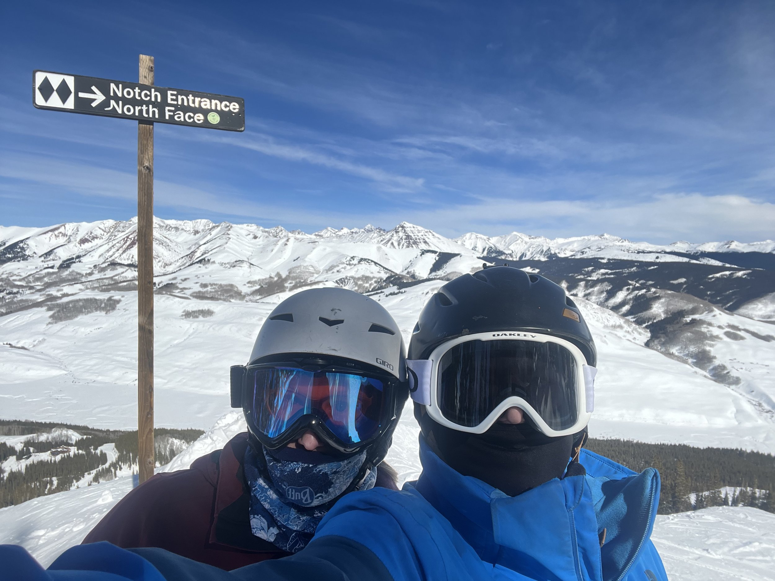 Two people wearing ski helmets, goggles, and winter clothing taking a selfie on a snow-covered mountain with a signpost and snow-capped mountains in the background.