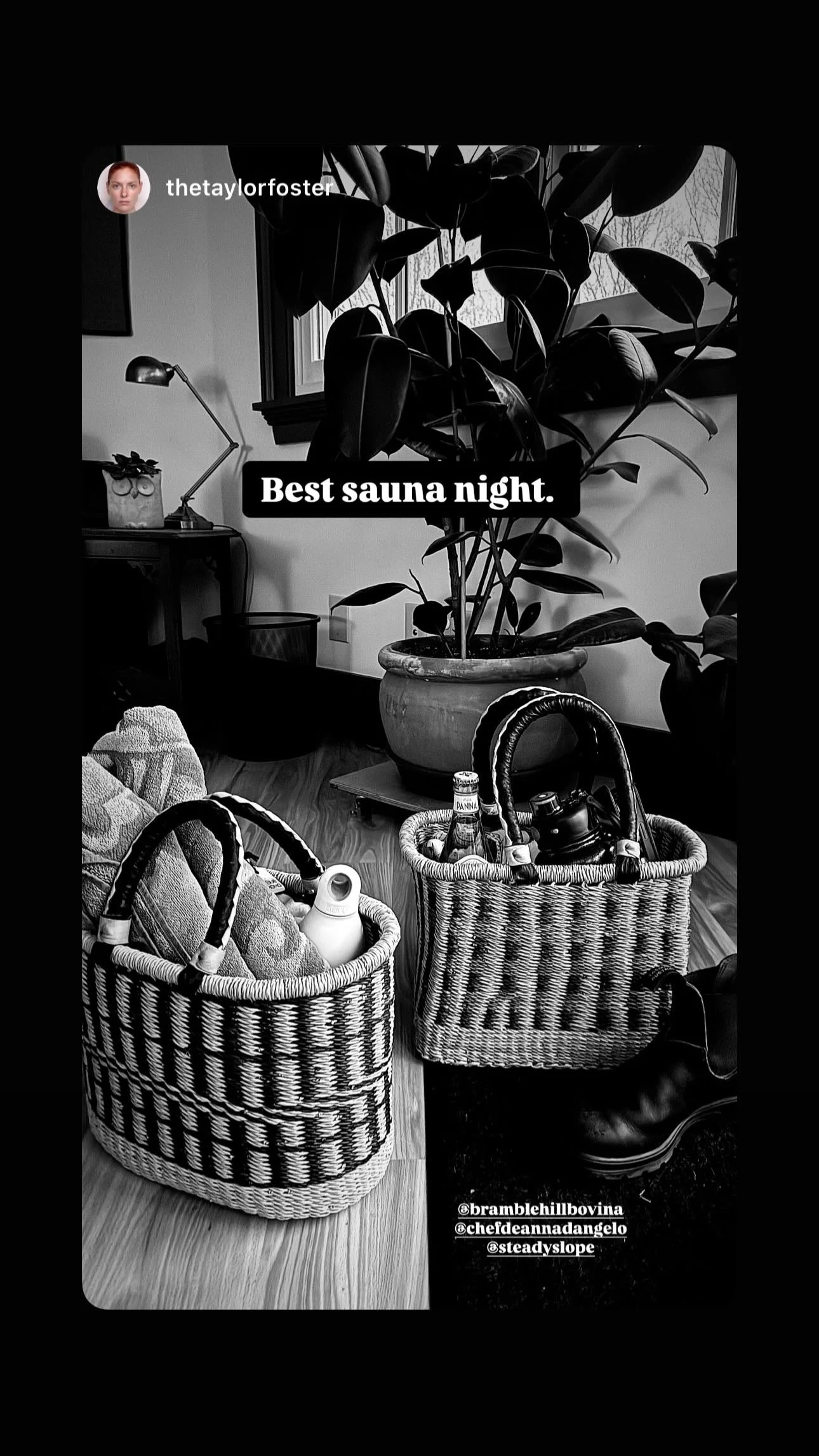 Black and white photo of a cozy indoor setting with plants, a desk lamp, and two woven baskets filled with items on a wooden floor, captioned "Best sauna night."