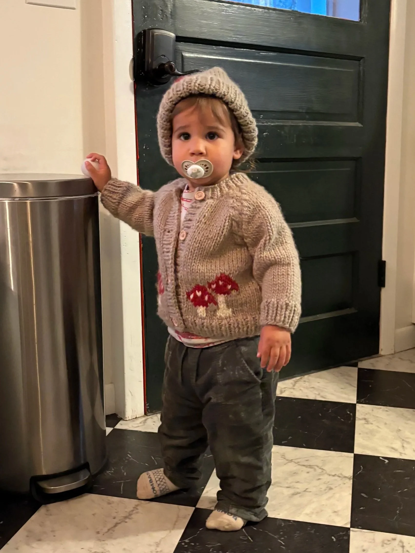 A young child with a pacifier in his mouth, wearing a beige knitted hat and cardigan with red mushroom patterns, dark pants, and socks, standing next to a trash can in front of a black door with a house interior background.