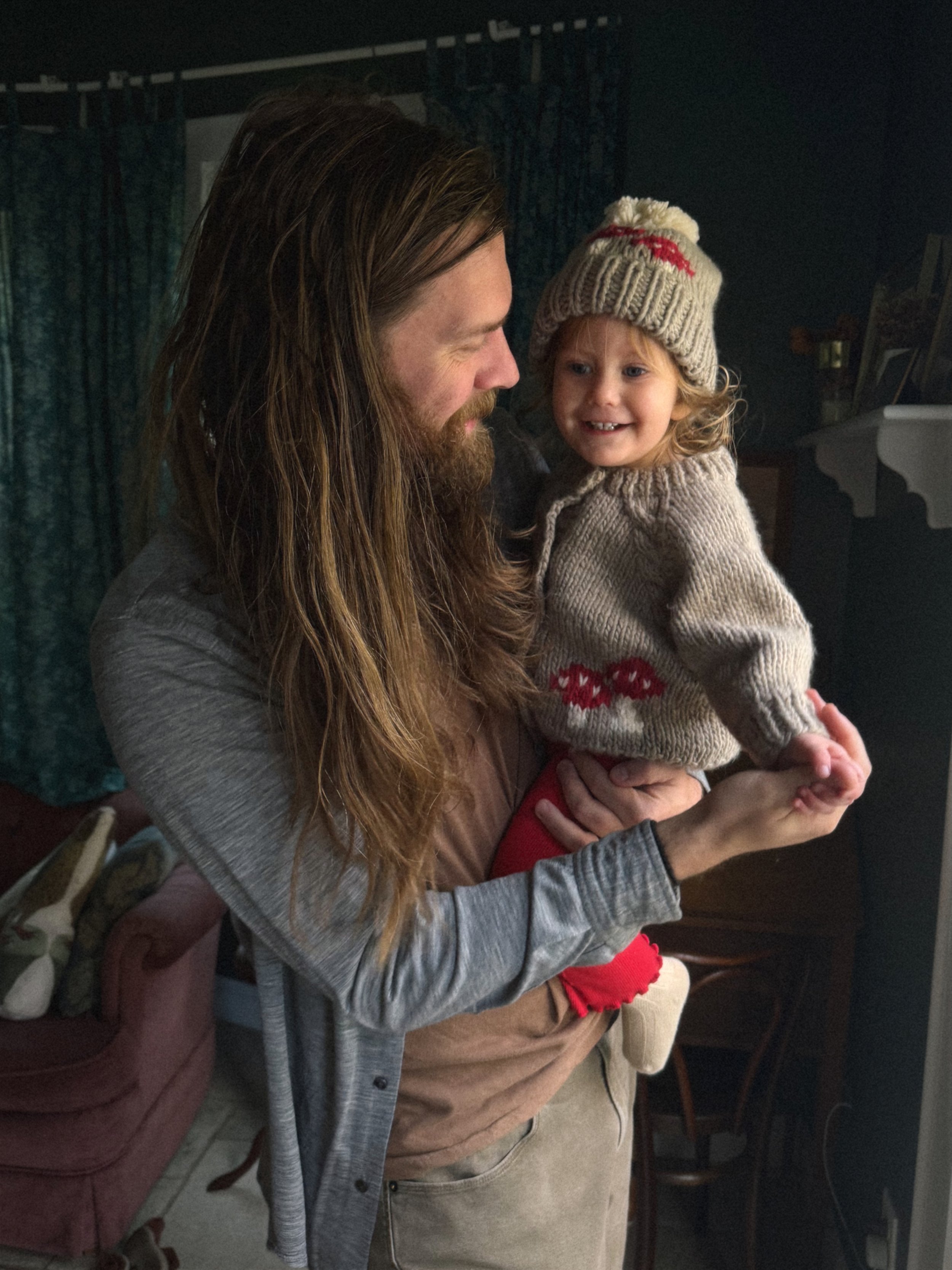 A man with long hair and a beard holding a young girl wearing a knit hat and sweater inside a house.