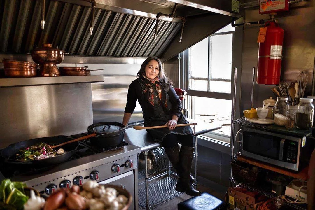 Woman sitting on a kitchen counter in a restaurant kitchen, holding a wooden spoon, surrounded by cookware, jars, and kitchen appliances.