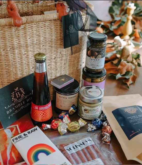 An assortment of food and drink items on a table, including a bottle of jarred syrup, jars of spreads, wrapped candies, a bag of chocolates, a box of chocolate, a package of meat sticks, and a gift basket in the background.