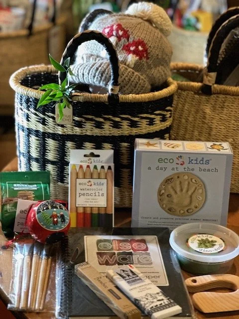 A collection of beach-related items and toys displayed on a table, including a woven basket containing a knitted hat with a red mushroom pattern, a set of watercolor pencils, wooden alphabet blocks, a package of eco-friendly craft supplies, and a sma