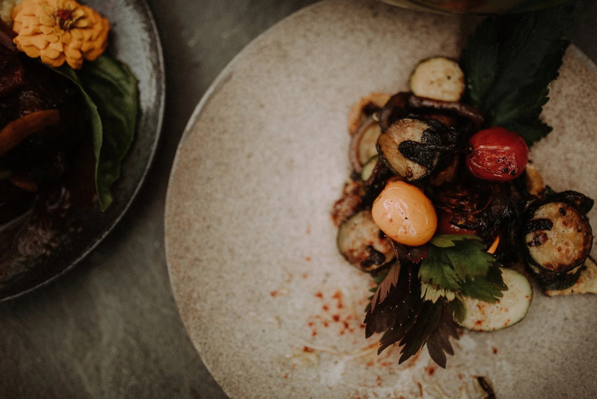 A plate of grilled vegetables including zucchini, cherry tomatoes, and possibly eggplant, garnished with herbs, on a textured white plate.