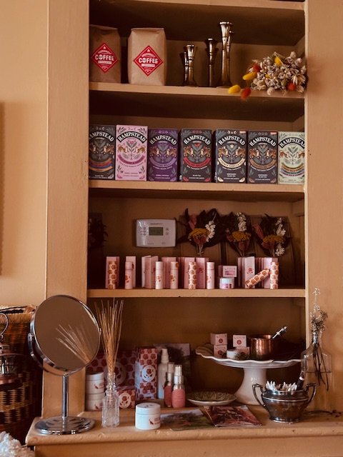 Coffee and tea supplies on a wooden shelf, including bags of coffee, boxes of tea, small decorative plants, a mirror, and various containers for liquids and spices.