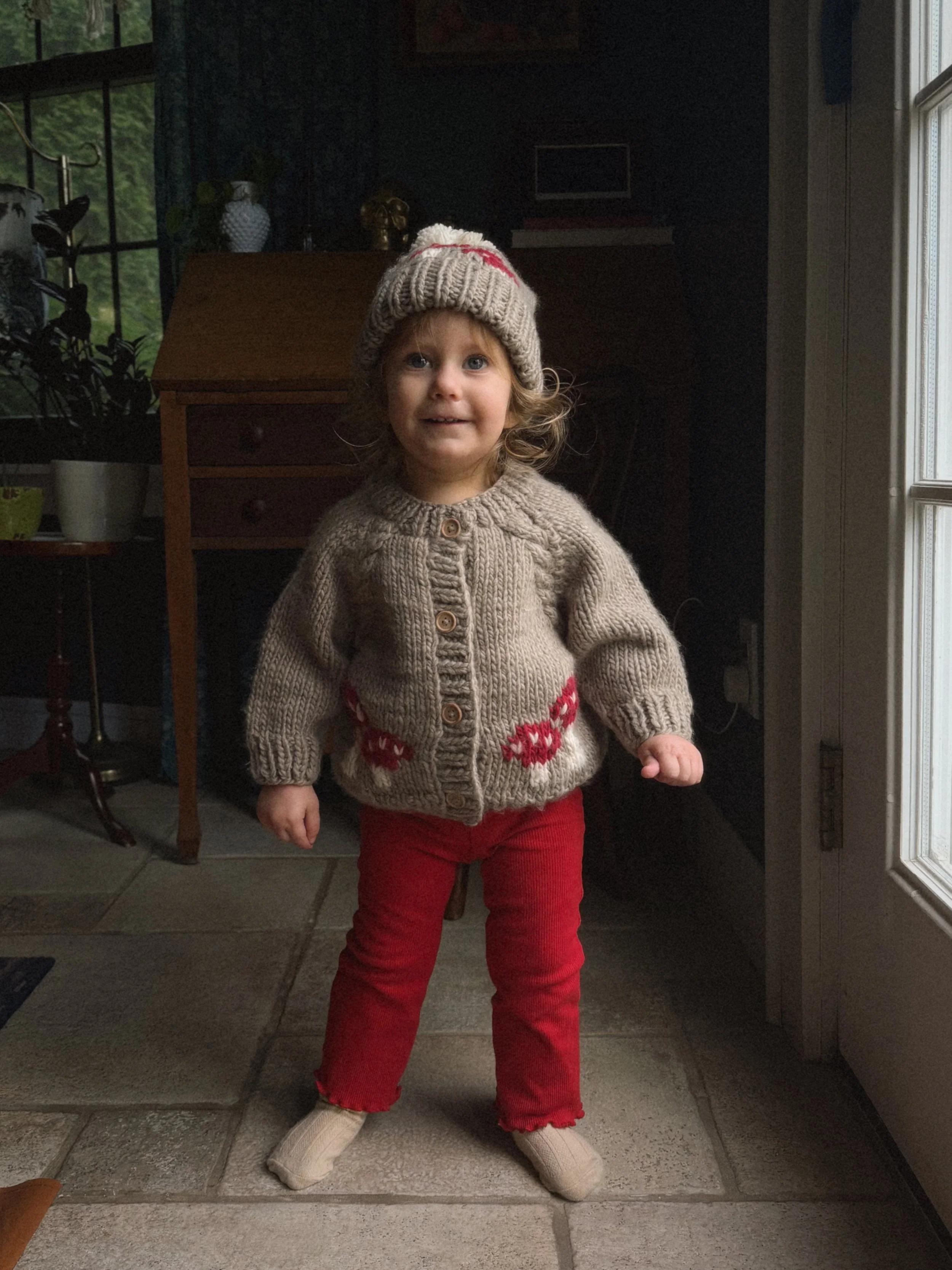 A young girl with curly hair, wearing a beige knit hat, a beige sweater with red reindeer, red pants, and beige socks, standing indoors near a glass door, smiling.
